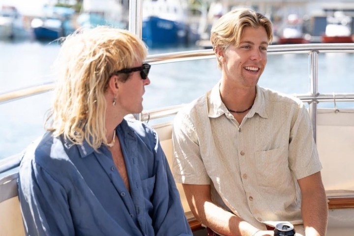 Two people smiling on a boat, one holding a drink, with a marina in the background.